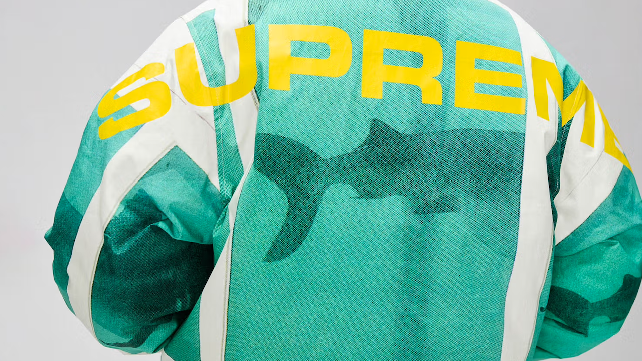 Una persona indossa una caratteristica giacca verde e bianca con un audace logo giallo "SUPREME" e un'accattivante sagoma di squalo sul retro, emblema dello stile streetwear audace.