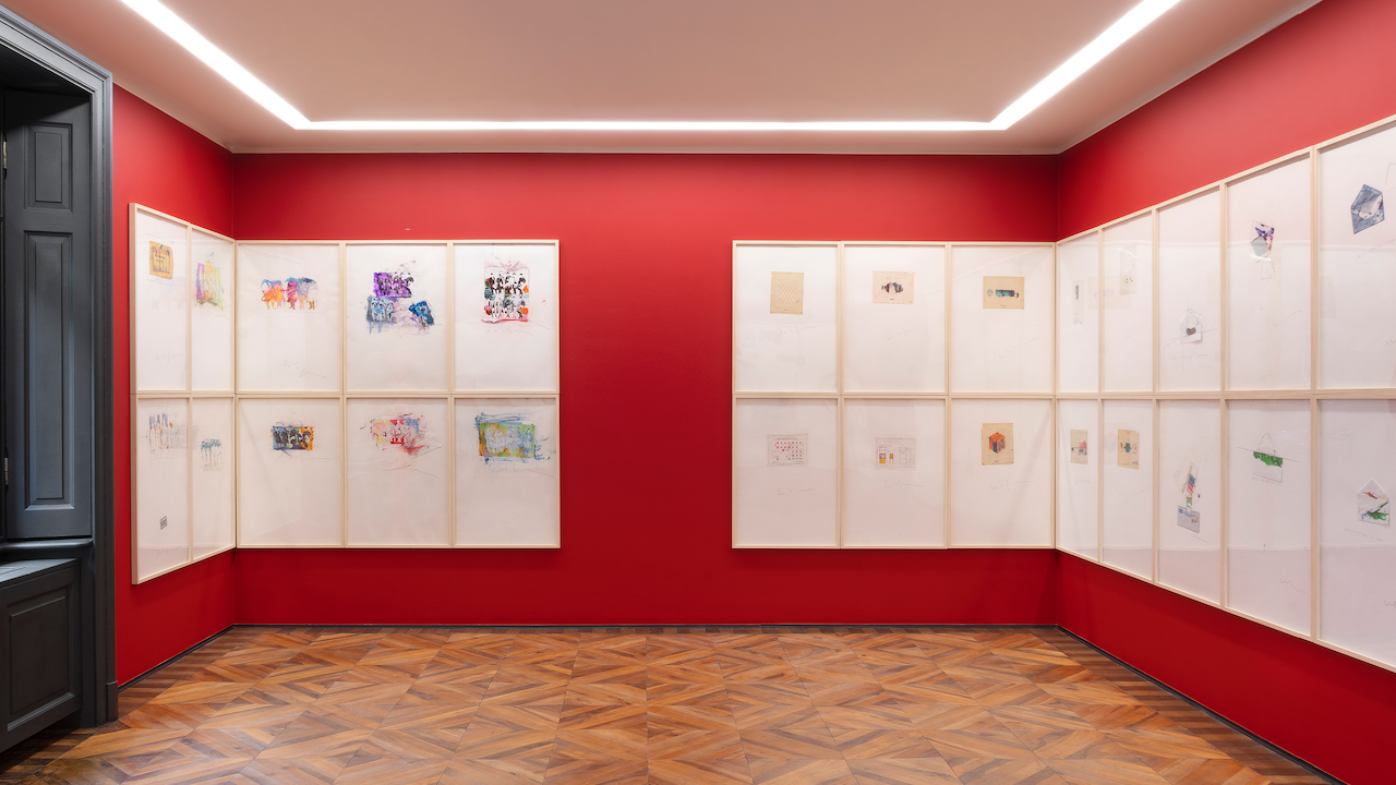 Una sala galleria con pareti rosse che espongono schizzi e dipinti incorniciati. Le opere d'arte sono disposte in una griglia su tre pareti. Il pavimento ha un motivo geometrico in legno e il soffitto è dotato di illuminazione da incasso.
