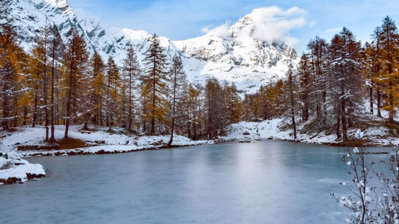 Un lago sereno e ghiacciato circondato da alberi innevati sotto un cielo azzurro e terso. Sullo sfondo, montagne innevate si ergono maestose, con nuvole sparse che coprono parzialmente le cime.