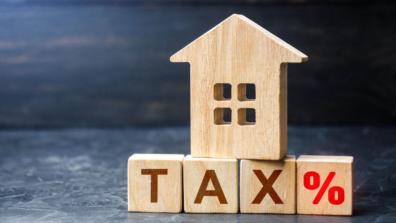 Un piccolo modello di casa in legno è appoggiato su blocchi, sempre di legno, che compongono la scritta "TAX" con un simbolo di percentuale, alludendo al tema delle tasse e delle agevolazioni casa. Lo sfondo, scuro e sfocato, attira l'attenzione sulla casa e sui blocchi.