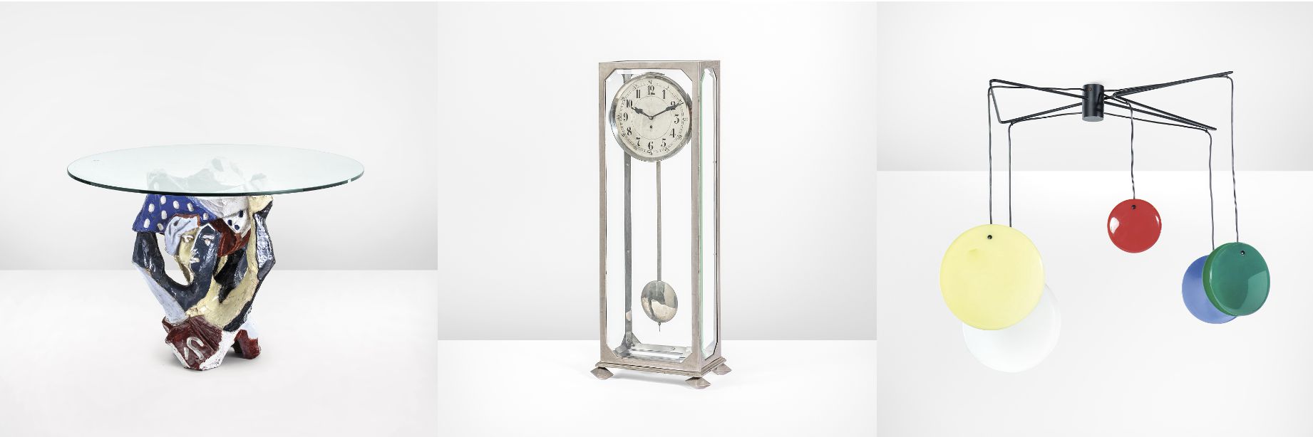 Un tavolo con piano in vetro e base astratta e colorata; un orologio da appoggio vintage con quadrante rotondo e pendolo; una moderna lampada da soffitto con quattro paralumi colorati a forma di disco nei colori rosso, giallo, verde e blu.