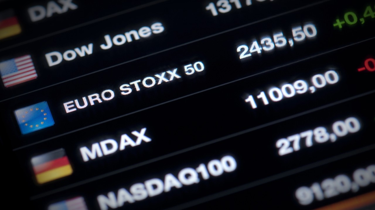 Primo piano di un display digitale che mostra vari valori di indici azionari. Lo schermo elenca Dow Jones a 24.355, EURO STOXX 50 a 11.009, MDAX a 2.778 e NASDAQ 100. Le bandiere rappresentano gli Stati Uniti, l'Unione Europea e la Germania accanto ai rispettivi indici.