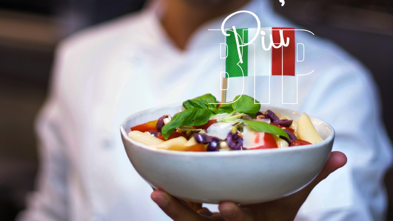 Uno chef in camice bianco che tiene in mano una ciotola di insalata di pasta guarnita con pomodorini, olive e basilico. La bandiera italiana e il testo sono sfocati sullo sfondo.