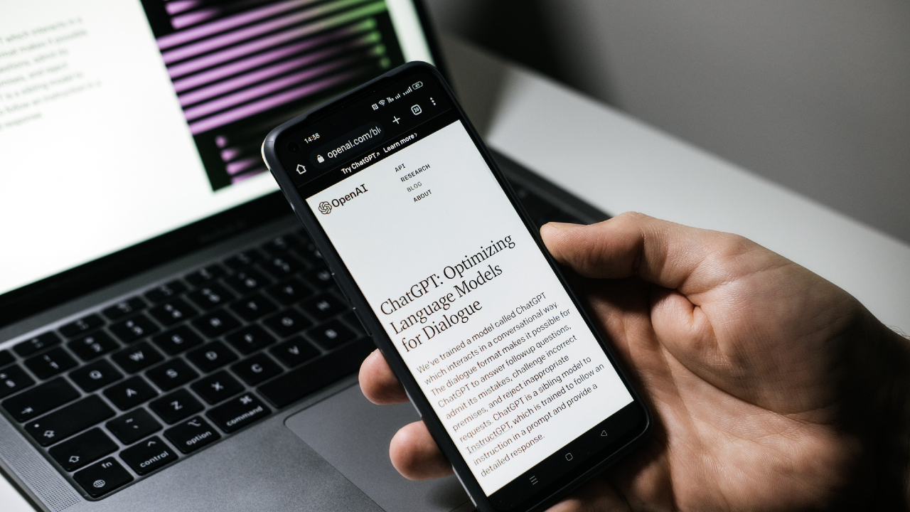 Una persona tiene in mano uno smartphone su cui è visualizzato un articolo su ChatGPT di OpenAI, con lo schermo sfocato di un laptop sullo sfondo.