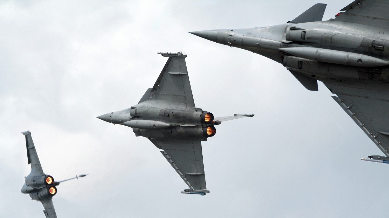 Tre jet da combattimento in volo si librano in un cielo nuvoloso. Due jet sono visti da dietro con i postbruciatori attivati, mentre il terzo jet è ripreso rivolto in avanti nell'angolo in alto a destra. La scena trasmette movimento dinamico e potenza.