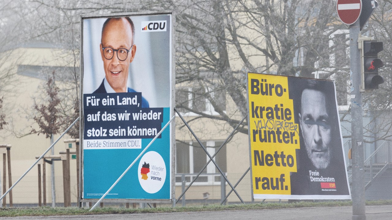 Due manifesti di campagna politica su una strada. Il manifesto a sinistra con un uomo sorridente promuove il partito CDU. Il manifesto a destra mostra il volto di un uomo dall'aria seria con un testo che sostiene la riduzione della burocrazia e l'aumento del reddito netto, promuovendo i Liberi Democratici.