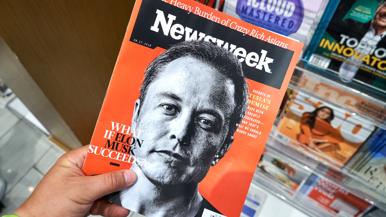 Una persona tiene in mano una rivista Newsweek con un ritratto in bianco e nero di un uomo su sfondo rosso. Il titolo di copertina recita: "E se Elon Musk avesse successo?". Sullo sfondo, sullo scaffale, sono visibili varie altre riviste e un articolo di OpenAI.