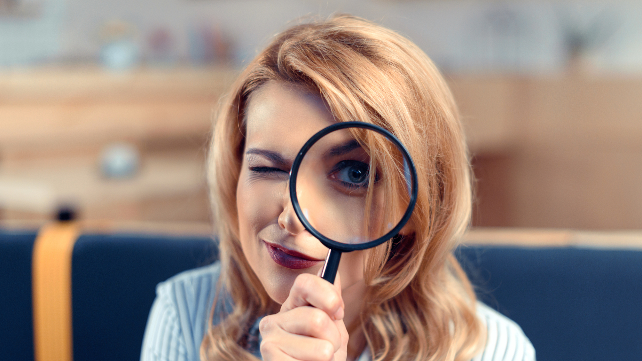 Una donna con i capelli biondi usa una lente di ingrandimento per ingrandire giocosamente un occhio mentre ammicca con l'altro. Nel mezzo di un'analisi di investimento eltif, è in un ambiente chiuso e lo sfondo leggermente sfocato crea un'atmosfera calda e spensierata.