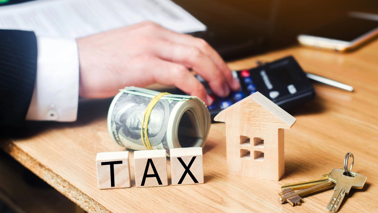 Una persona usa una calcolatrice su una scrivania. Nelle vicinanze ci sono ricevute di bollette, un rotolo di banconote da un dollaro Usa, lettere maiuscole in legno che compongono la parola "Tax", una piccola casa in legno e un mazzo di chiavi. Il riferimento va ai calcoli per la trasformazione da holding immobiliari a società semplice con imposta 8% nel 2026.