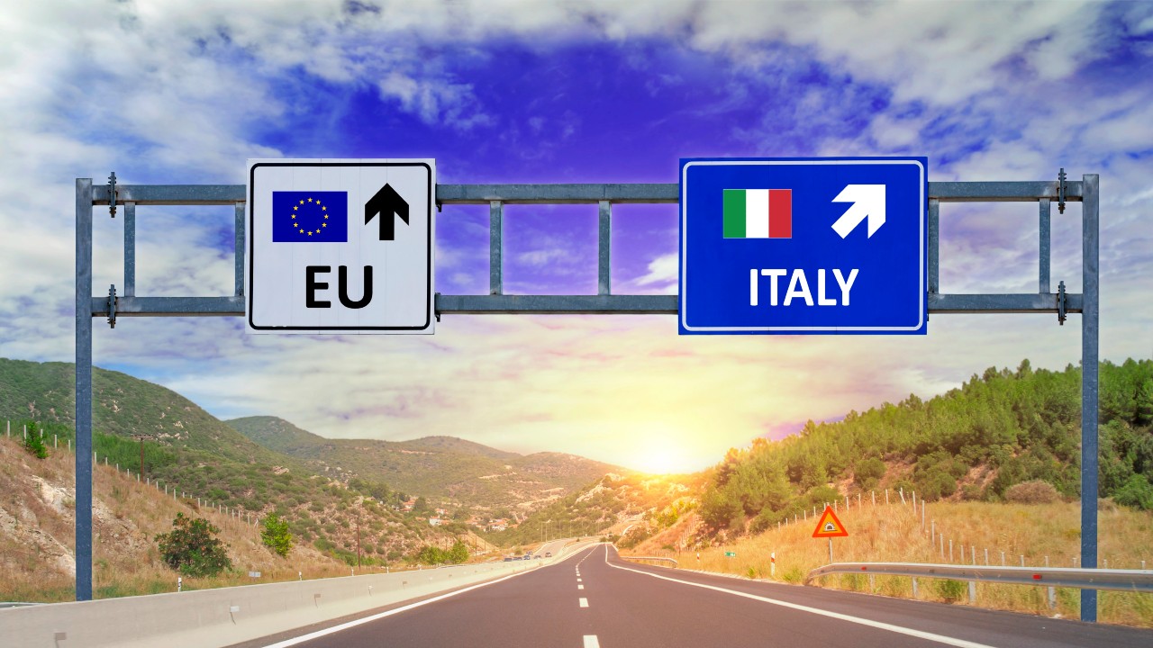 Due segnali stradali autostradali con frecce indicano le direzioni da prendere: uno mostra la bandiera dell'Ue con una freccia rivolta verso l'alto, mentre un altro mostra la bandiera italiana che punta a destra. Il riferimento va al tema dell’esterovestizione societaria.