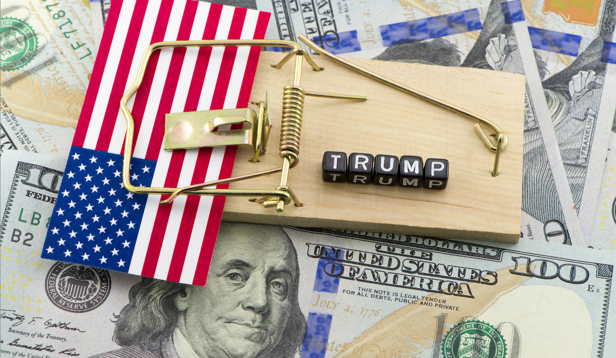 Una trappola per topi con una piccola bandiera americana e blocchi di lettere che compongono la scritta "TRUMP" è posizionata in cima a una pila di banconote da 100 dollari. La bandiera è agganciata accanto alle lettere sulla base di legno della trappola.