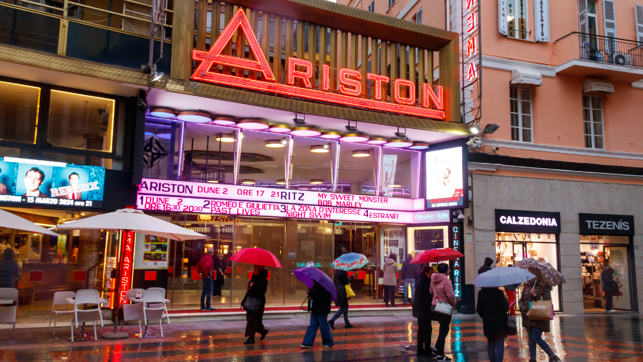 Persone con ombrelli camminano davanti al Teatro Ariston di Sanremo, illuminato di notte. L'insegna al neon mostra le date dei prossimi spettacoli. La zona è bagnata dalla pioggia e i negozi circostanti hanno insegne luminose.