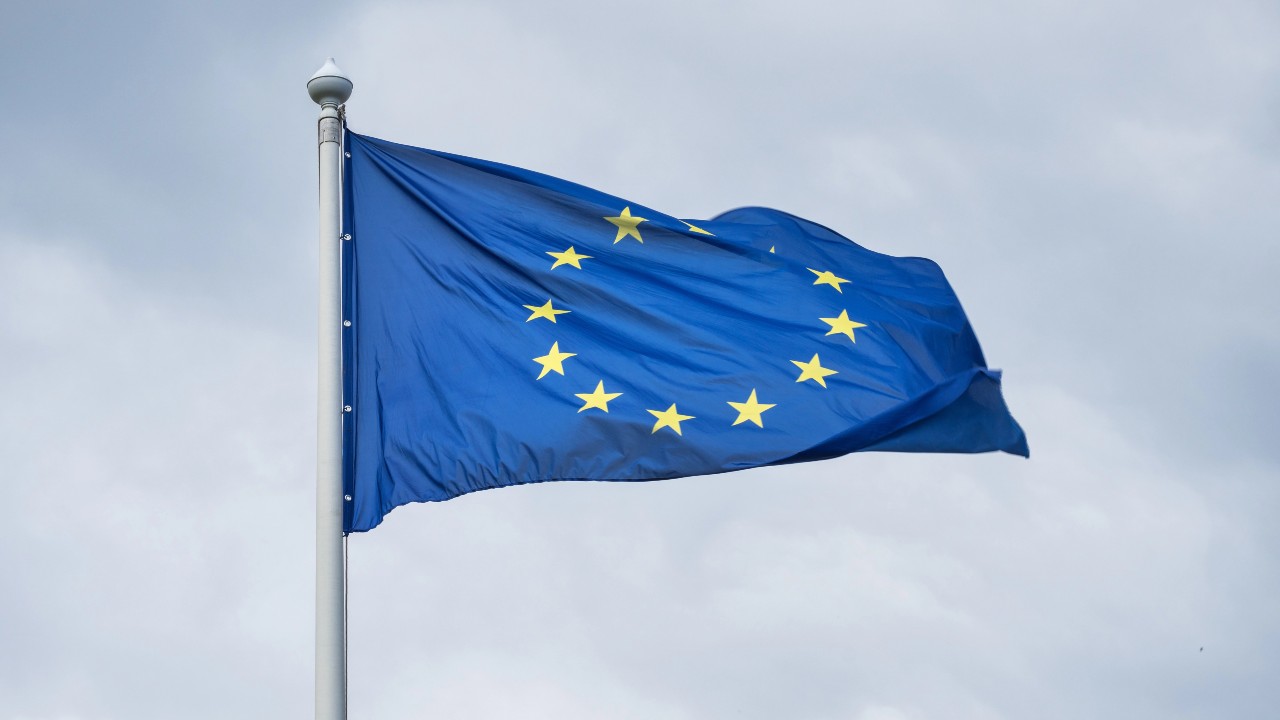 L'immagine mostra la bandiera dell'Unione Europea, caratterizzata da un cerchio di dodici stelle dorate su uno sfondo blu. La bandiera sventola su un palo contro un cielo nuvoloso.
