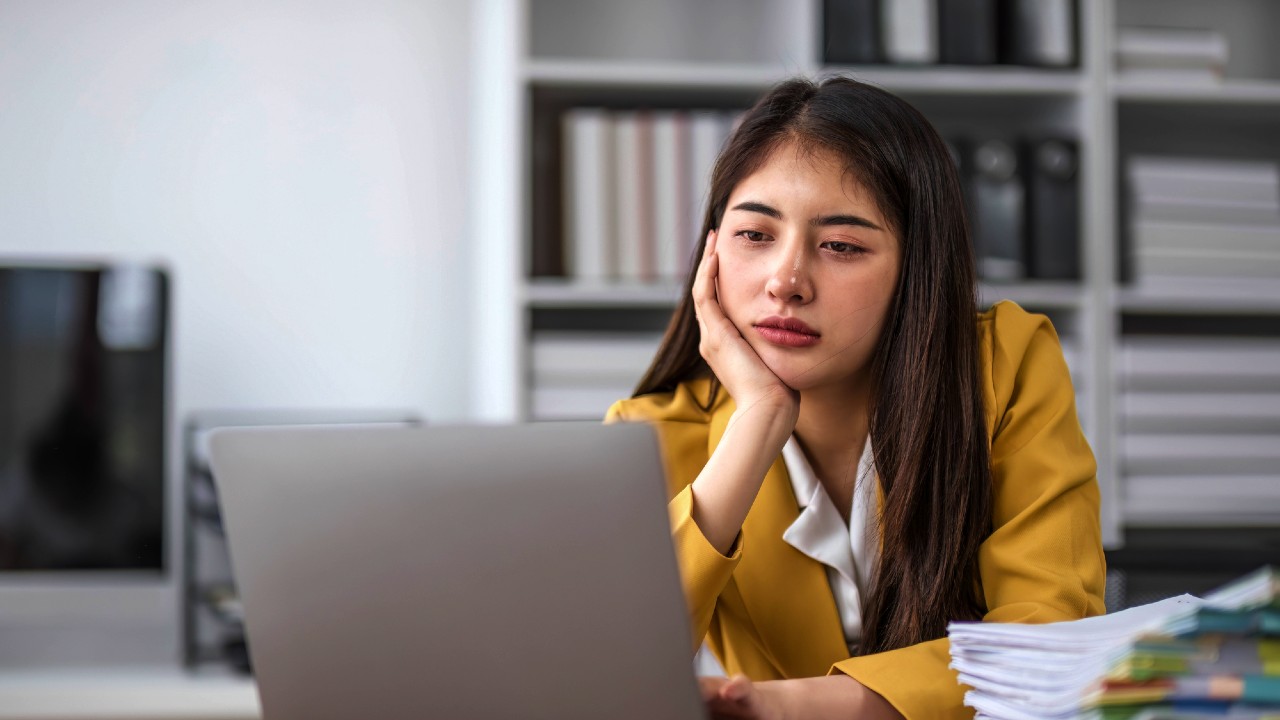 Una donna con un blazer giallo è seduta a una scrivania, appoggia il viso sulla mano mentre guarda lo schermo di un computer portatile. Sembra pensierosa o preoccupata. Sullo sfondo, c'è una libreria piena di libri e raccoglitori.