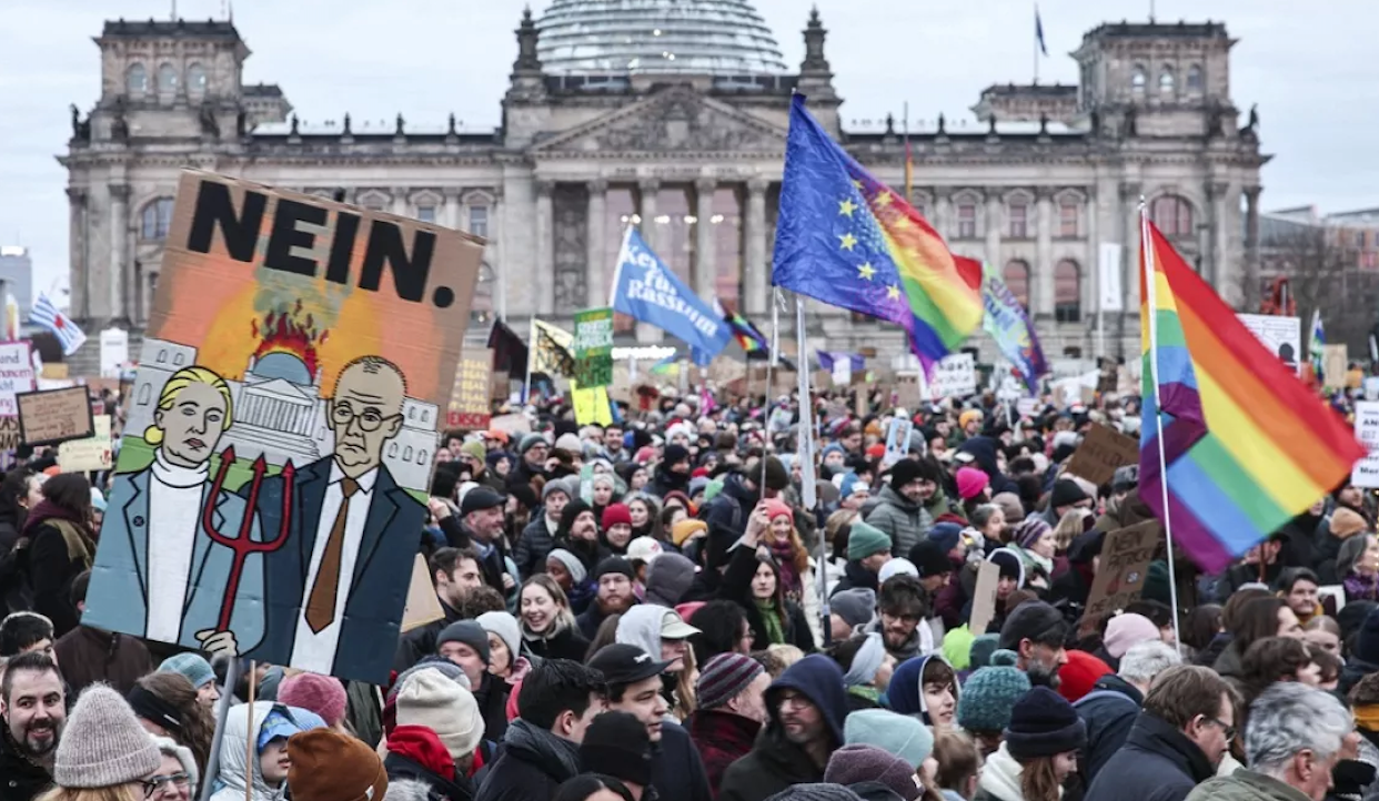 Una grande folla si raduna a Berlino davanti al parlamento, con cartelli e bandiere, tra cui bandiere arcobaleno e dell'UE. Un cartello in evidenza raffigura due figure con la parola "NEIN". L'atmosfera è vivace e varia, con molti partecipanti ben coperti per il freddo.