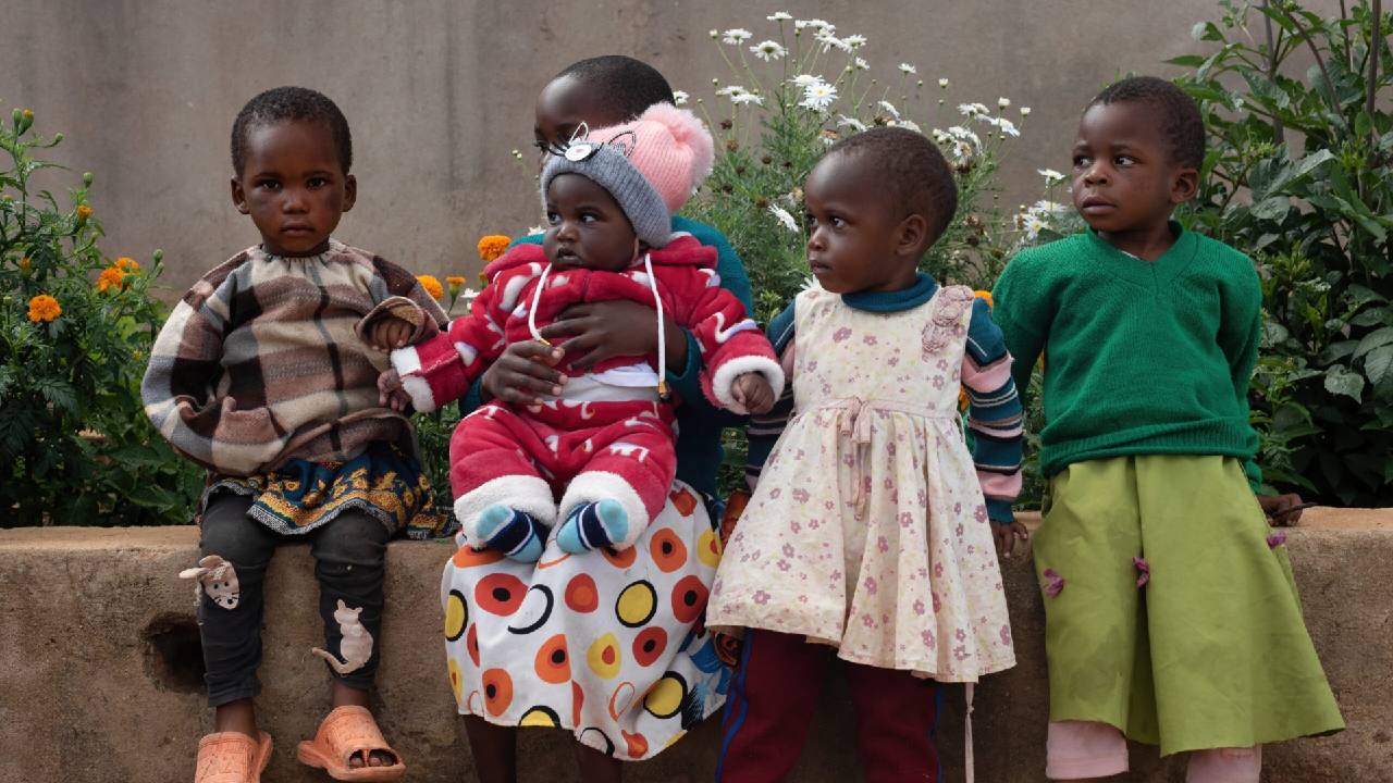 Cinque bambini piccoli siedono insieme su un muretto basso in Tanzania. Uno tiene in braccio un neonato vestito di rosso. Sono circondati da verde e fiori vivaci, e indossano abiti colorati. Lo sfondo è un muro semplice, che riflette il fascino locale.
