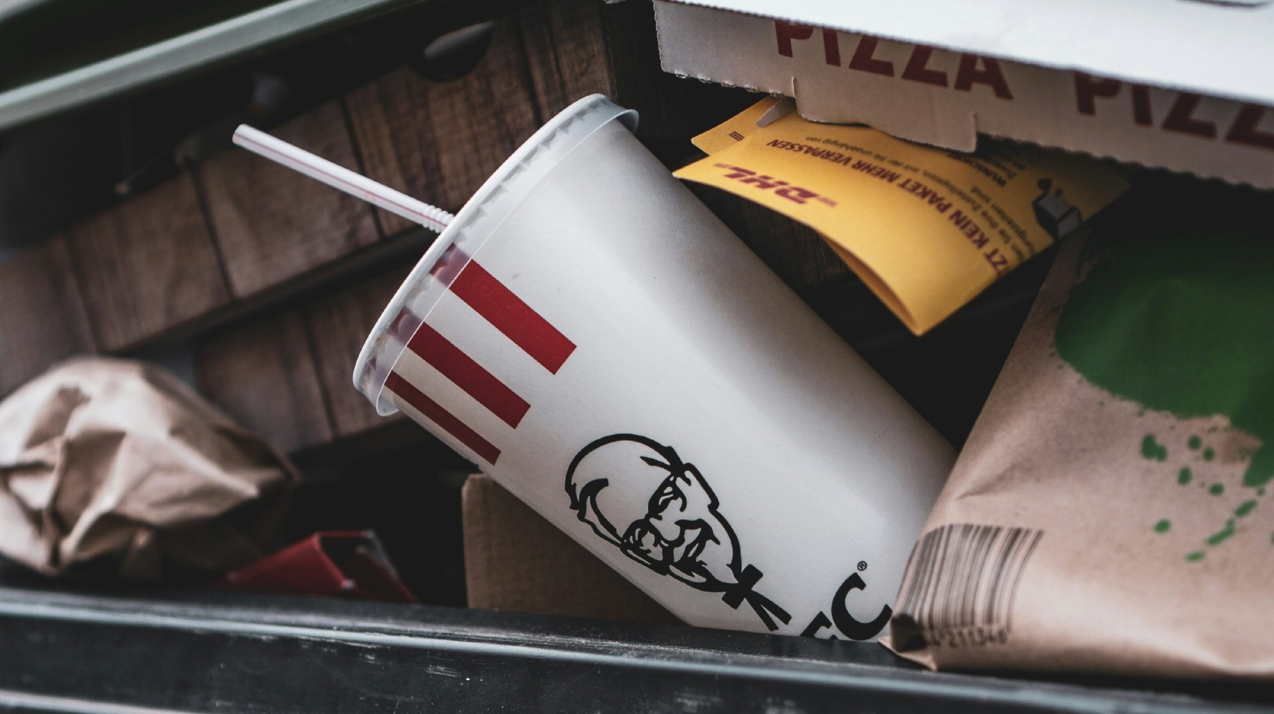 Un primo piano di un bidone della spazzatura pieno di rifiuti di fast food. Gli oggetti visibili includono un bicchiere di soda schiacciato di Carmignac Investimenti con strisce rosse e un logo riconoscibile, scatole di pizza e sacchetti di carta marrone. La scena trasmette un senso di disordine e spreco urbano.