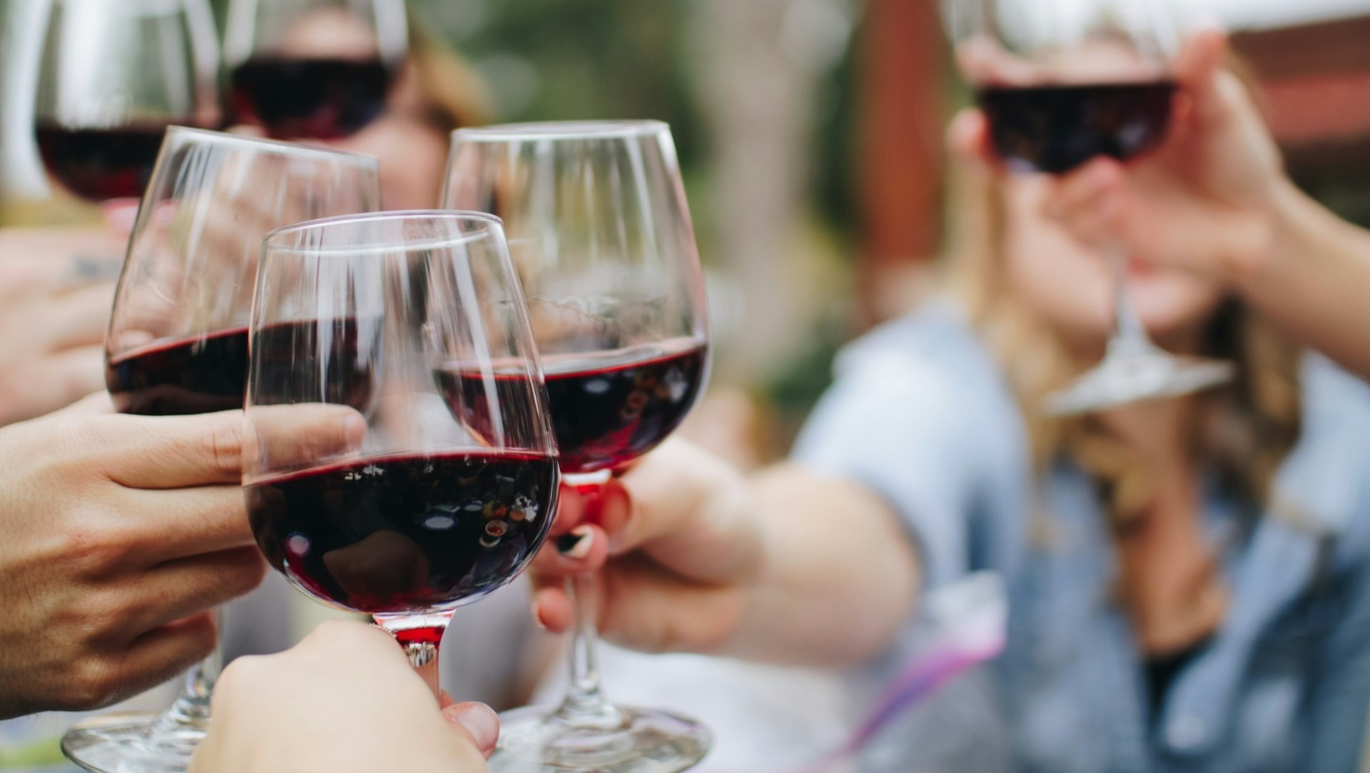 Primo piano di diverse mani che brindano con bicchieri di vino rosso all'aperto. Lo sfondo è sfocato, focalizzato sui bicchieri di vino e sul gesto celebrativo.