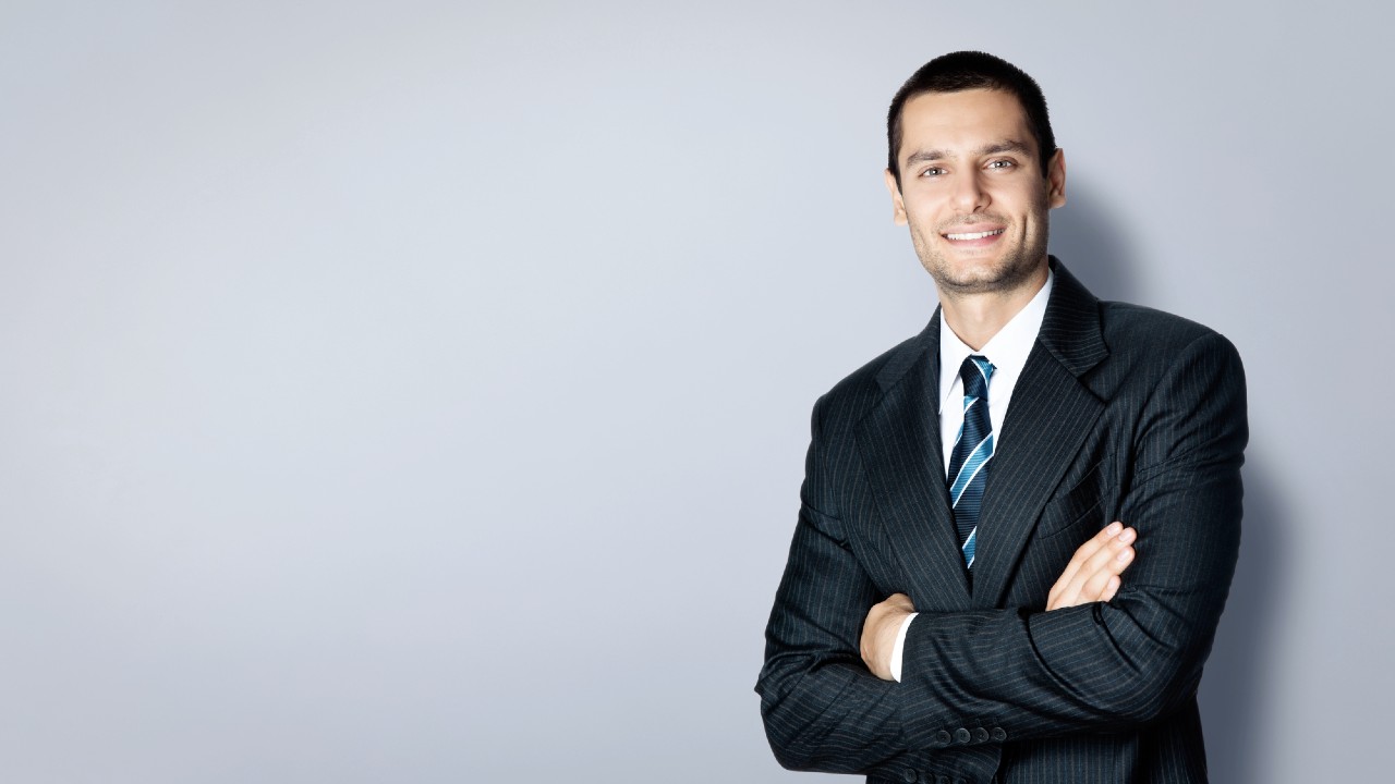 Un uomo in giacca e cravatta a righe blu sta in piedi con sicurezza su uno sfondo grigio chiaro, con le braccia incrociate e sorridente.