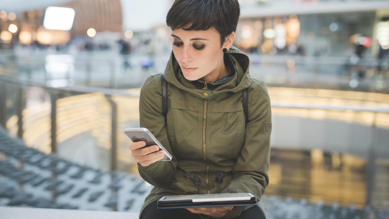 Una persona con i capelli corti e una giacca verde è seduta in un interno, con in mano uno smartphone e un tablet, immersa nella tecnologia. Sullo sfondo di un vivace centro commerciale con luci brillanti e un'energia vibrante, la scena cattura l'essenza della vita moderna che aspira alla longevità.
