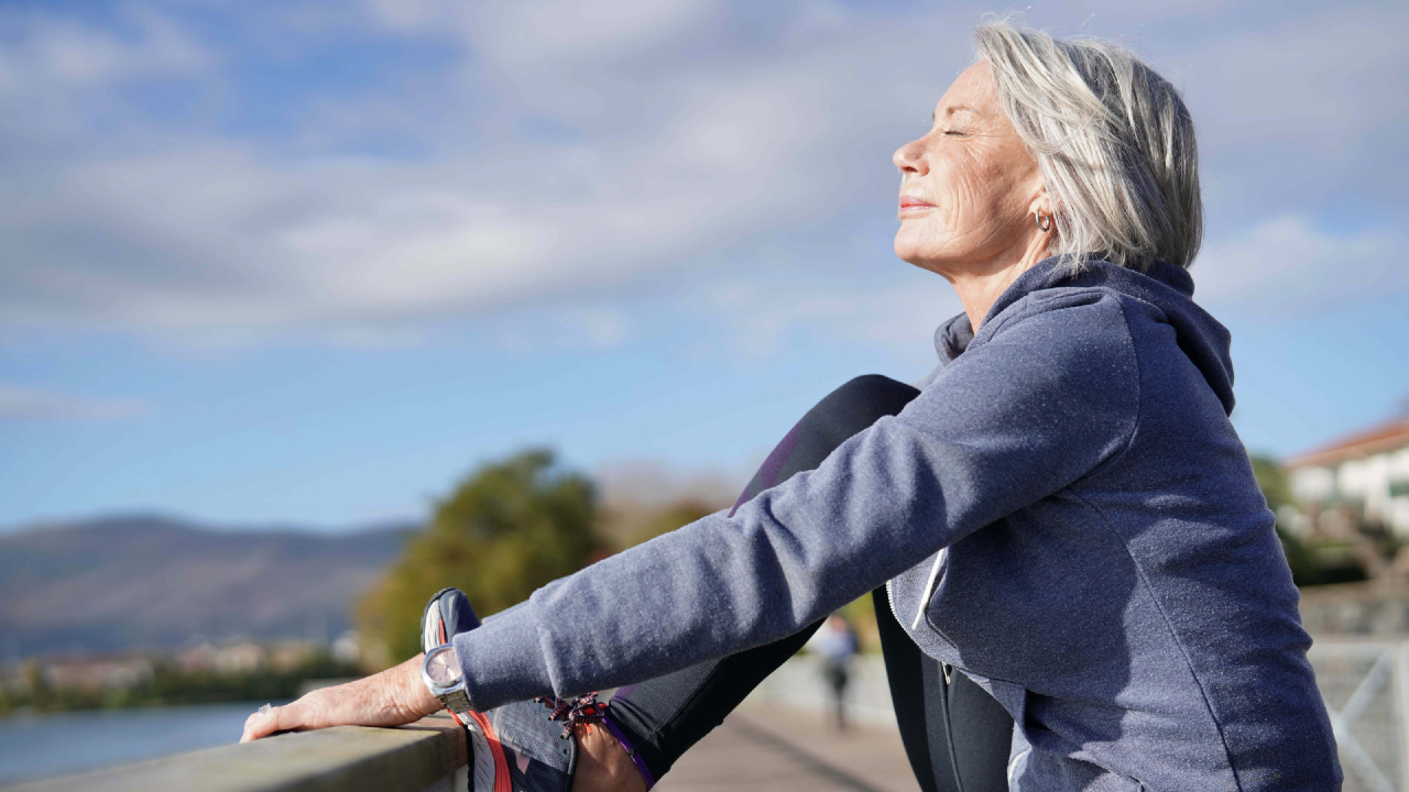 Una donna anziana con i capelli argentati, in felpa con cappuccio e abbigliamento sportivo, si allunga, incarnando uno spirito di longevità, con un ginocchio piegato su una ringhiera. Ha gli occhi chiusi e il viso inclinato verso il sole. Il cielo nuvoloso e lo sfondo naturale sfocato di alberi e colline completano la scena serena.