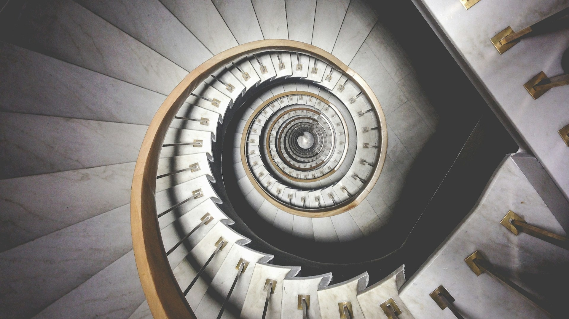 Vista aerea di una scala a chiocciola con gradini in marmo e corrimano in legno. La scala forma un motivo ipnotico che converge al centro, con accenti dorati lungo i corrimano. La prospettiva crea un effetto visivo sorprendente.