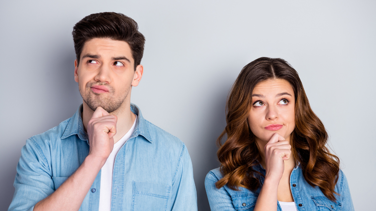 Un uomo e una donna sono in piedi uno accanto all'altro su uno sfondo grigio chiaro, riflettendo su possibilità diverse. Entrambi hanno le mani sul mento e indossano camicie casual in denim blu. La donna ha lunghi capelli ondulati, mentre l'uomo sfoggia capelli corti e acconciati.