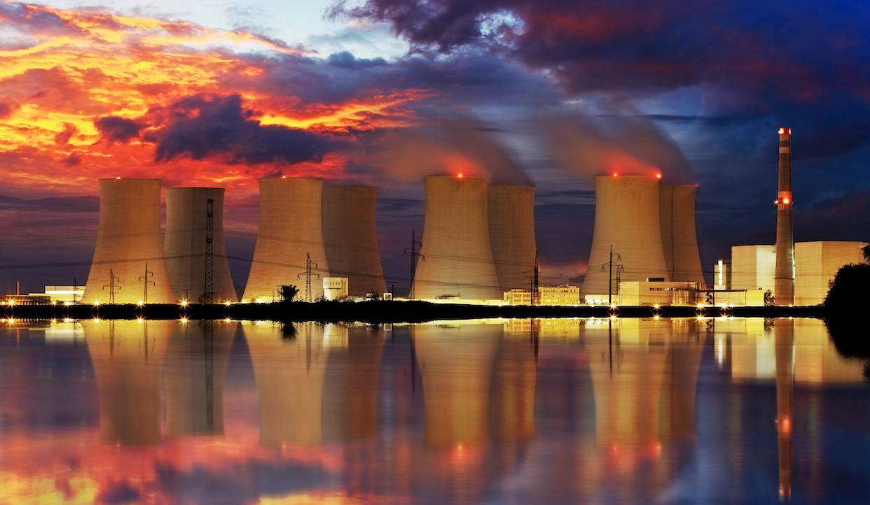 Una centrale nucleare con torri di raffreddamento che emettono vapore, illuminata contro un vibrante cielo al tramonto. La scena si riflette su uno specchio d'acqua in primo piano, creando un'immagine speculare simmetrica della centrale e delle drammatiche nuvole soprastanti.