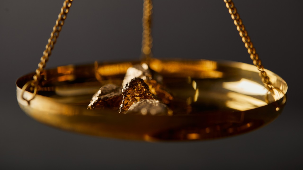 Un primo piano di una bilancia dorata con diverse pepite d'oro grezzo poste su un lato. La bilancia è sospesa da catene e lo sfondo è scuro, evidenziando la superficie lucida e la consistenza dell'oro.