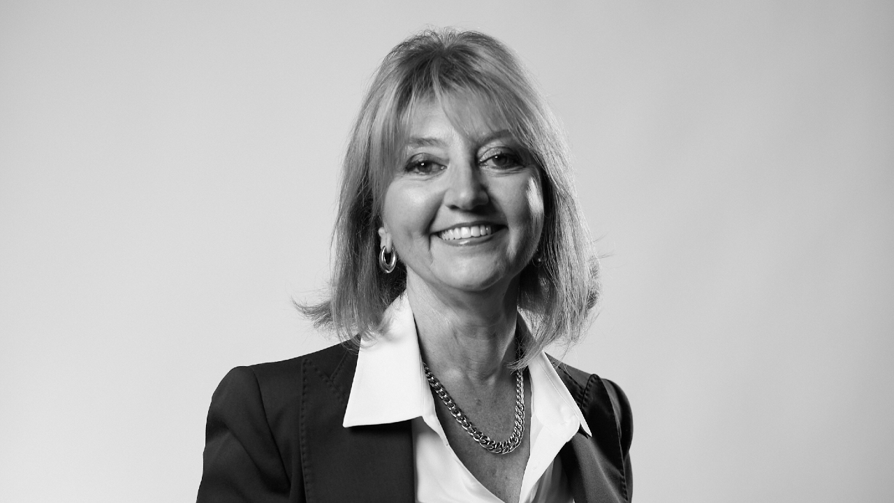 Ritratto in bianco e nero di una donna con i capelli lunghi fino alle spalle, sorridente. Indossa un blazer scuro sopra una camicetta chiara con una collana di corna spessa. Lo sfondo è una tonalità semplice e chiara, che ricorda lo stile classico Pellegrini.