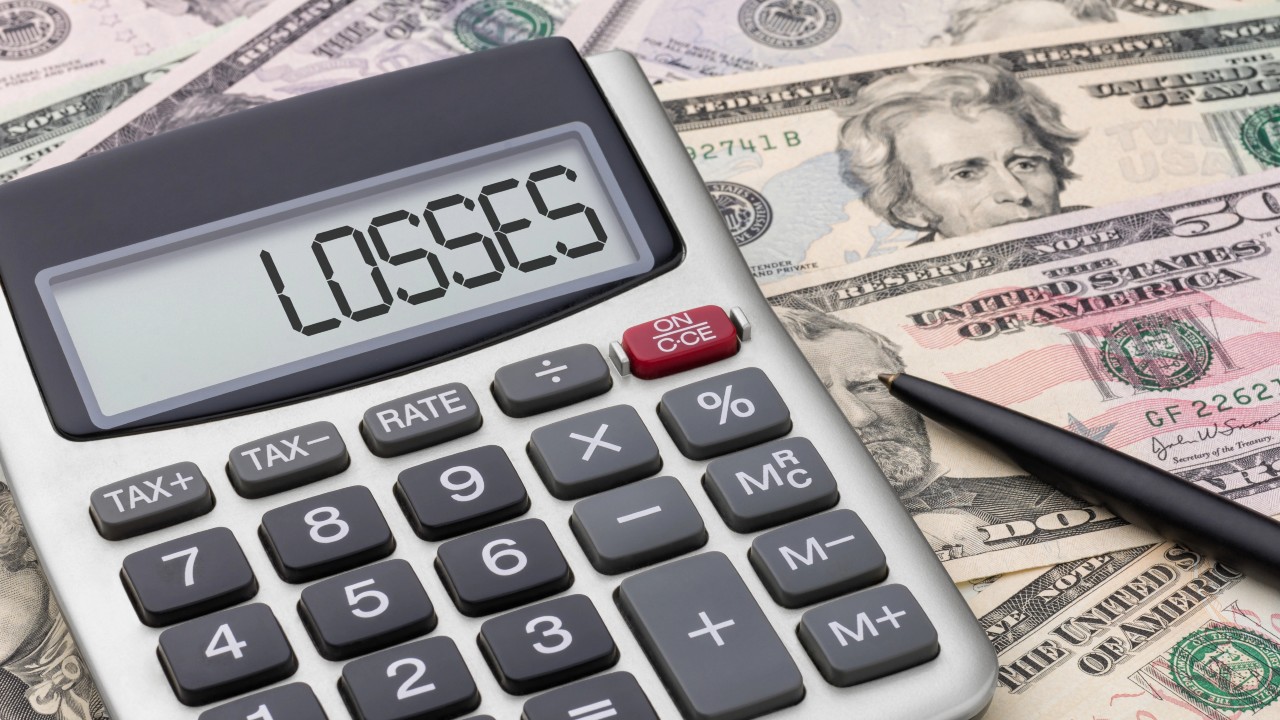 Un primo piano di una calcolatrice mostra la parola "PERDITE", circondata da banconote da un dollaro statunitense e da una penna nera, a simboleggiare calcoli finanziari e possibili perdite finanziarie.