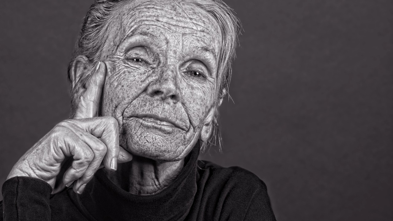 Una persona anziana con profonde rughe d'espressione sul viso guarda direttamente la telecamera. Appoggia il mento sulla mano e sembra pensierosa. Lo sfondo è un grigio semplice. L'immagine è in bianco e nero.