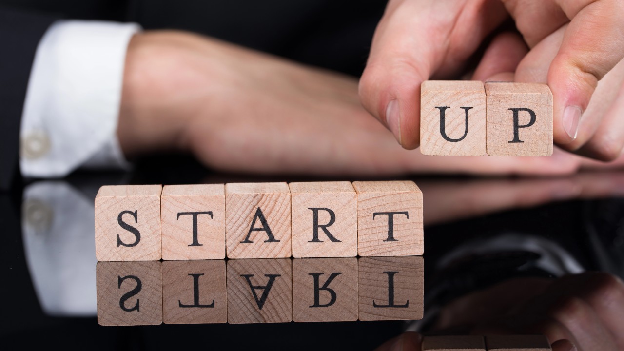 Primo piano di blocchi di legno che compongono la scritta "STARTUP" su una superficie riflettente. Una mano tiene alcuni blocchi, mentre l’altra è distesa, illudendo a idee innovative su startup e pmi.