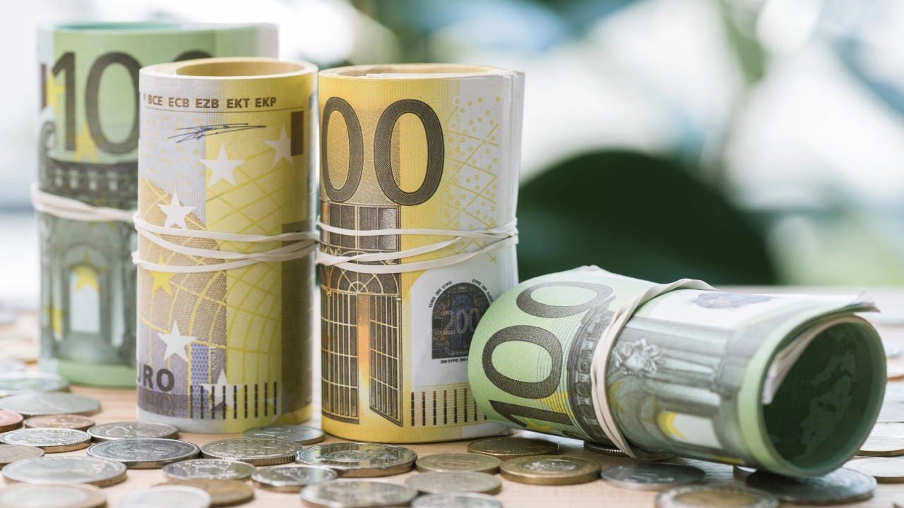 Delle banconote di denaro contante in euro sono arrotolate con elastici e circondate da varie monete su una superficie piana. Le denominazioni visibili includono 10, 50 e 100 euro. Lo sfondo è sfocato, con tenui tonalità di verde.