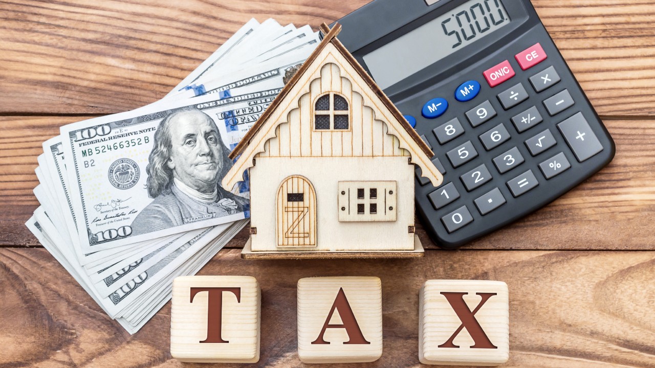 Un piccolo modello di casa in legno è posto tra una pila di banconote da cento dollari e una calcolatrice. Sotto di loro, dei blocchi di legno compongono la parola "tassa". Il riferimento va alla Participation exemption (Pex) e alle plusvalenze.