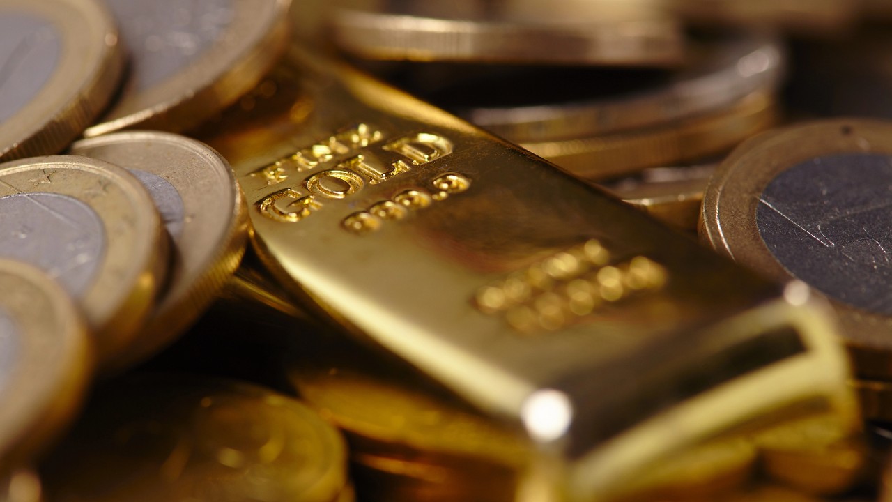 Primo piano di una barra d'oro lucida incisa con la scritta "FINE GOLD" circondata da numerose monete d'oro. La barra e le monete sono impilate, riflettendo la luce per evidenziare la loro superficie metallica.