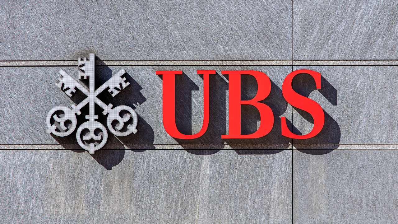 L'immagine mostra l'iconico logo UBS su un muro di pietra grigia, con tre chiavi incrociate in metallo accanto alle lettere rosse in grassetto "UBS".