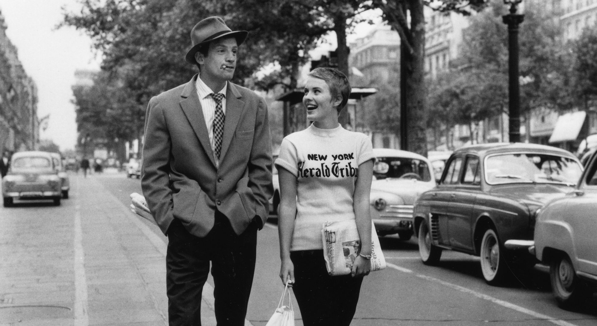Una foto in bianco e nero di un uomo in giacca e cravatta e di una donna con una maglietta del "New York Herald Tribune" che camminano felici lungo una strada di Parigi. Auto d'epoca sono parcheggiate lungo il marciapiede e alberi costeggiano la strada trafficata sullo sfondo.