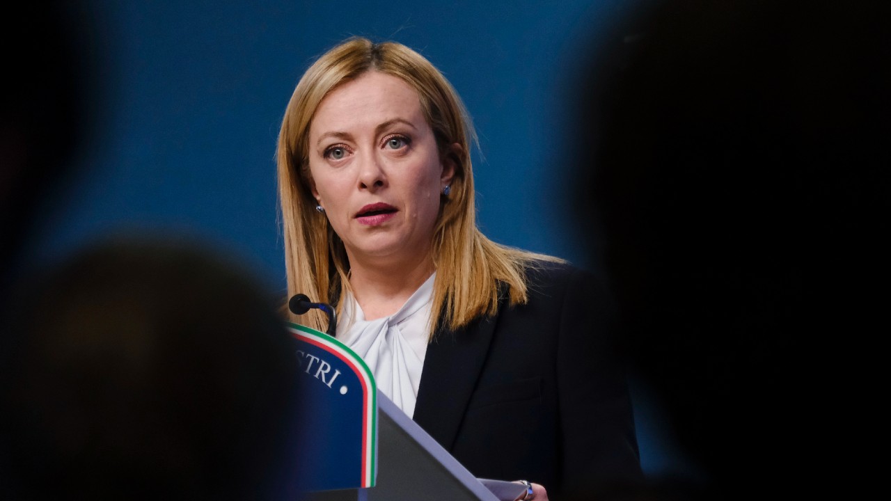 Una persona con lunghi capelli biondi sta parlando su un podio con un microfono. Indossa un blazer nero sopra una maglietta bianca e lo sfondo è blu. In primo piano si vedono sagome sfocate di altre persone.