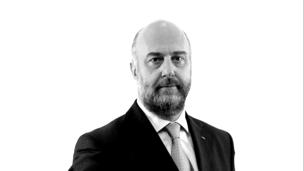 Un'immagine in bianco e nero di un uomo calvo con la barba, che indossa un completo e una cravatta. Lo sfondo è bianco e lui guarda direttamente la telecamera con un'espressione neutra.