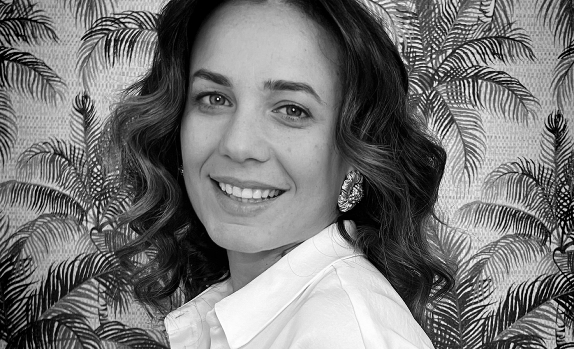 Donna con capelli mossi e sorridente, che indossa una camicetta bianca e grandi orecchini, in posa su uno sfondo con motivi a foglie di palma. Immagine in bianco e nero.