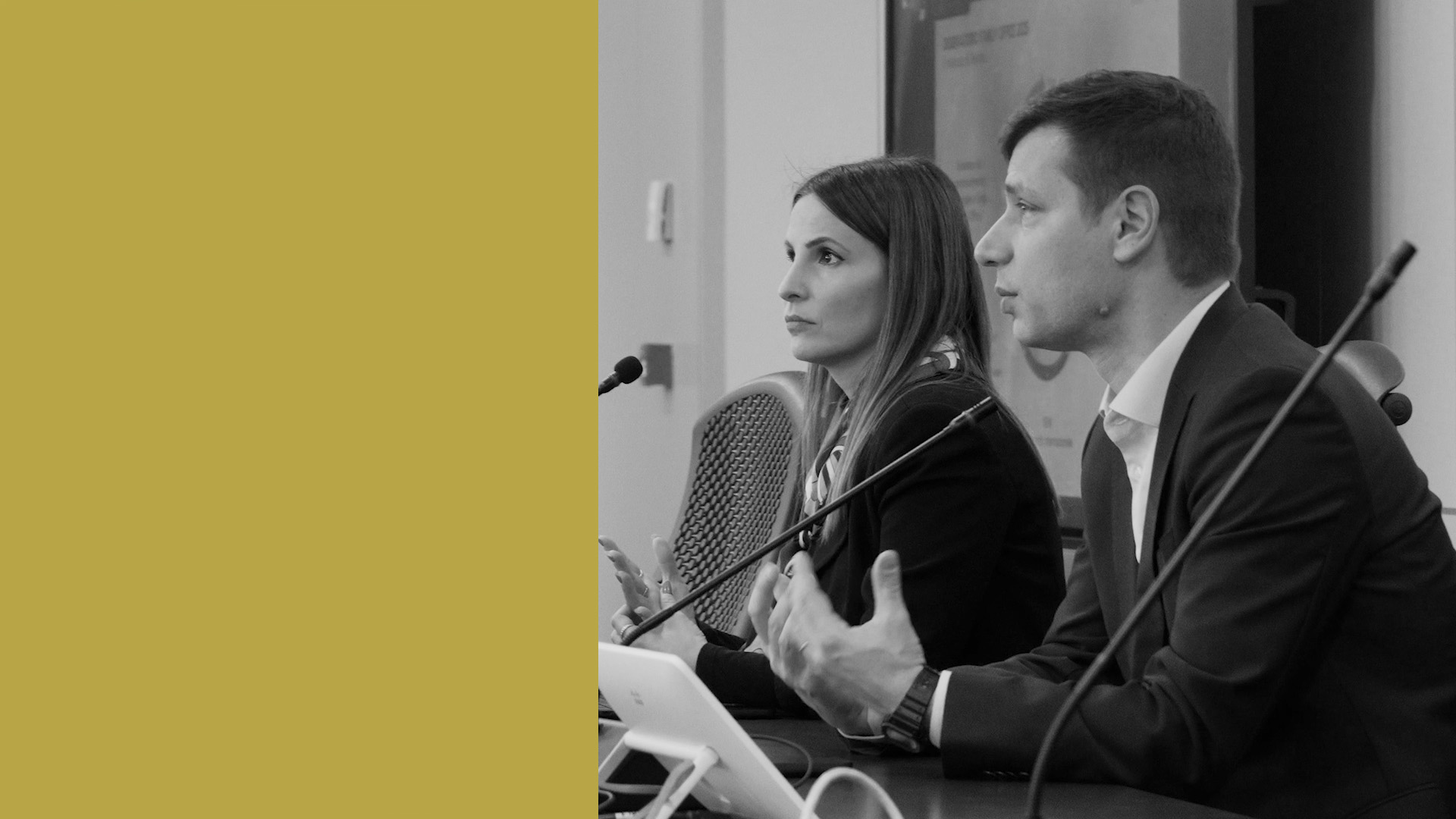 Un'immagine in bianco e nero di un uomo e una donna, Emanuela Rondi, in abiti da lavoro che parlano nei microfoni a un tavolo da conferenza. L'uomo è Josip Kotlar e gesticola con le mani e il tavolo contiene diversi dispositivi. Una grande area gialla riempie il lato sinistro dell'immagine.