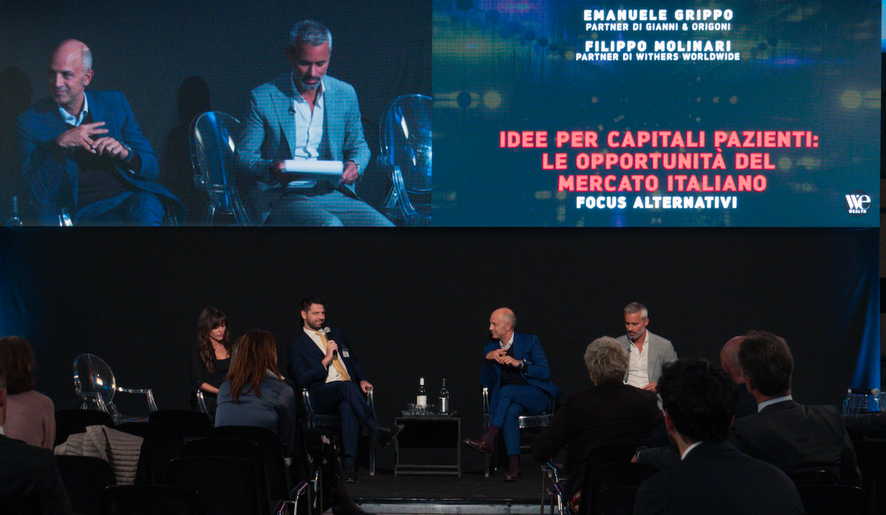 Quattro persone sono sedute su un palco per una tavola rotonda. Dietro di loro, un grande schermo mostra un testo sulle opportunità nel mercato italiano con i nomi di due partner di studi legali. L'ambiente è professionale, con un pubblico concentrato.
