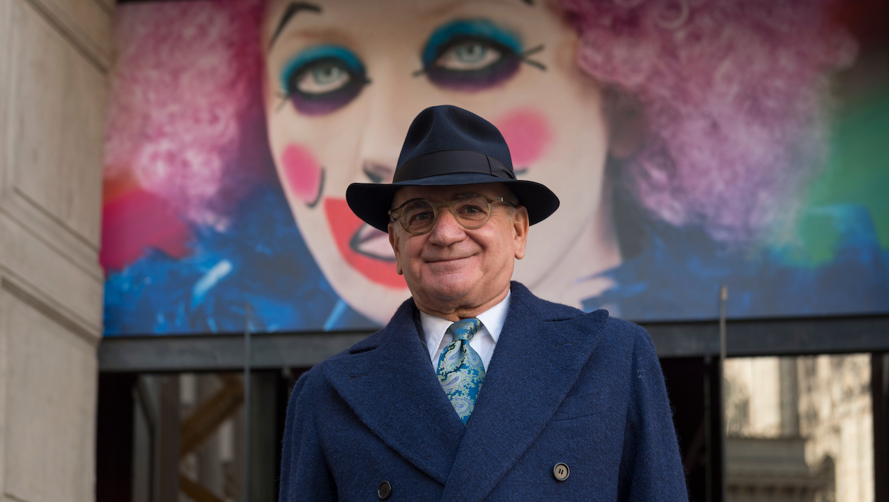 Una persona sorridente con un cappotto e un cappello blu scuro è in piedi davanti a un grande e colorato murale di clown con capelli rosa, occhi esagerati e guance rosee. Lo sfondo mostra parte di un edificio.