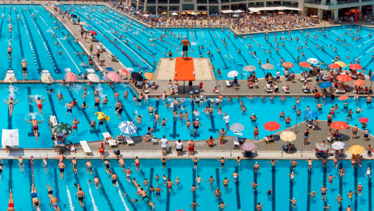 Vista aerea di un'affollata area di piscina pubblica con numerosi nuotatori e bagnanti. Diverse grandi piscine sono circondate da ombrelloni colorati e lettini prendisole. Un bagnino è in piedi su una piattaforma rialzata al centro, a sorvegliare la scena.