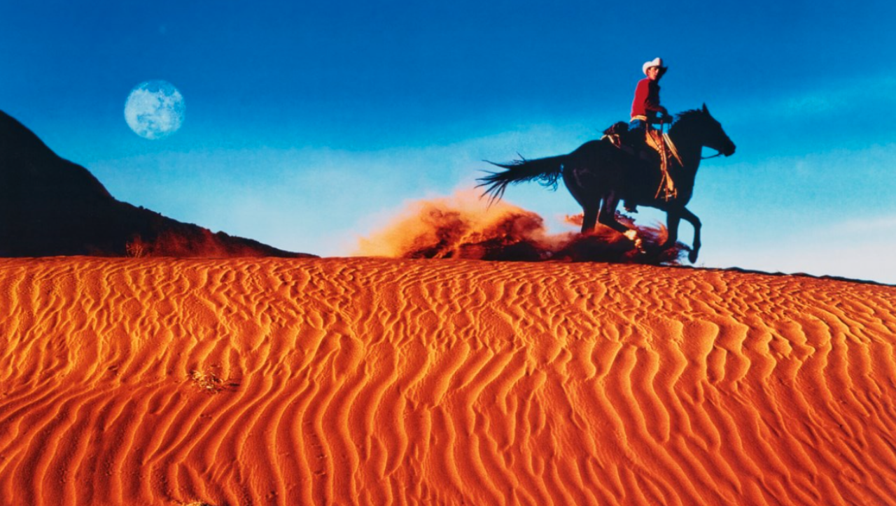 Una persona che indossa un cappello da cowboy cavalca un cavallo attraverso dune di sabbia arancione increspate sotto un cielo azzurro brillante. La luna è visibile sul lato sinistro del cielo, aggiungendo un tocco surreale alla vibrante scena desertica.