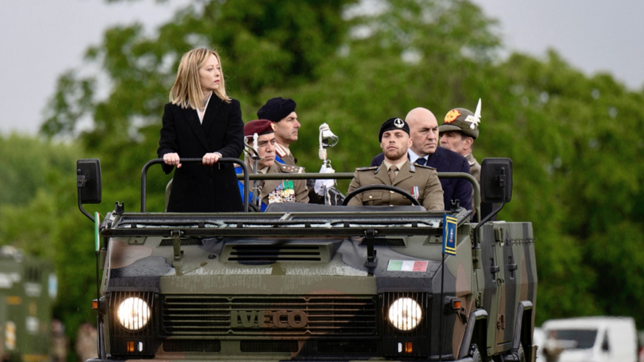 Una donna è in piedi su una jeep militare, accompagnata da diversi uomini in uniforme. Il veicolo ha una bandiera italiana e il gruppo sembra partecipare a un evento formale. Sullo sfondo si vedono degli alberi.