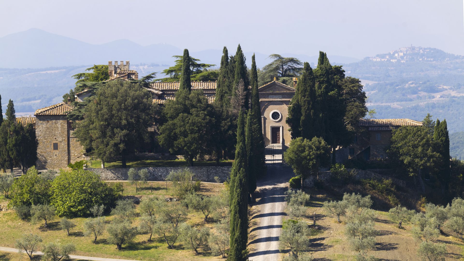 Villa in pietra con tetto in tegole circondata da alti cipressi e uliveti in cima a una collina. Un sentiero conduce all'ingresso. Colline ondulate e una città lontana sono visibili sotto un cielo azzurro e terso.