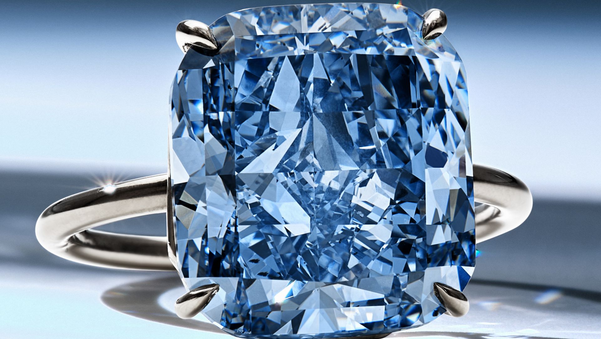 Un primo piano di un anello d'argento con un grande diamante blu dal taglio rettangolare, con sfaccettature che riflettono la luce. Lo sfondo è un delicato gradiente di blu e bianco.