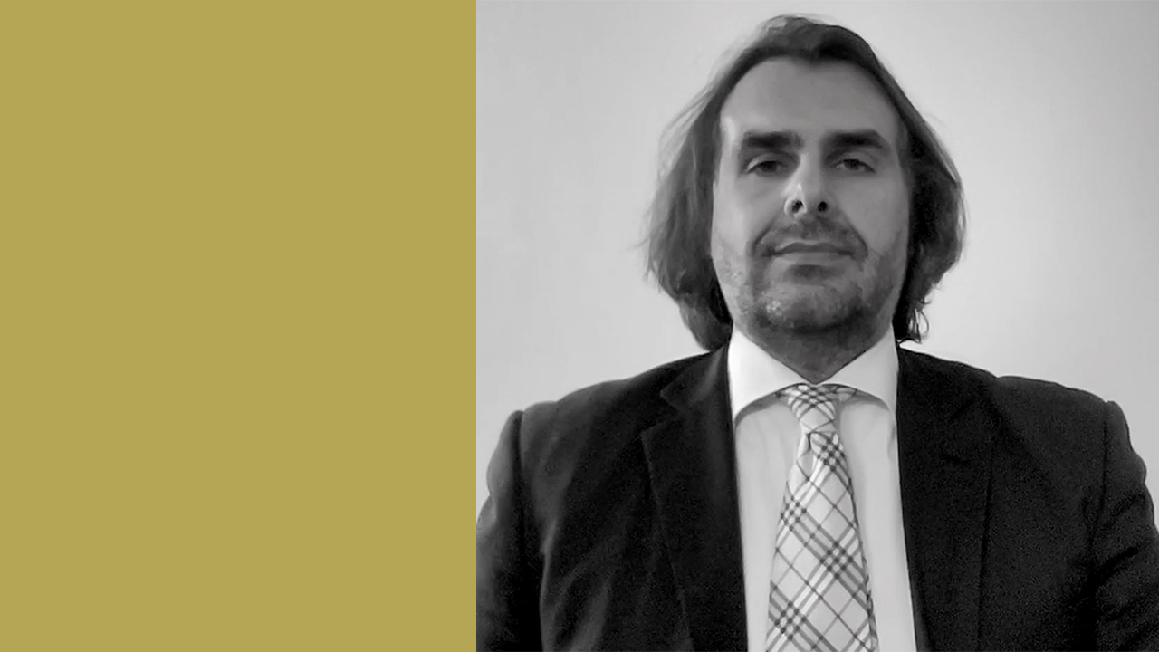 Un uomo con i capelli lunghi fino alle spalle e la barba, ovvero Gianluca Tonato di Endevo, indossa un abito e una cravatta scozzese, è raffigurato su uno sfondo semplice con una striscia verticale color senape a sinistra. L'immagine è in bianco e nero.